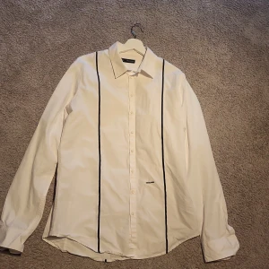 Vit skjorta från Dsquared2 med svarta ränder - Vit skjorta från Dsquared2 med svarta vertikala ränder framtill och broderad logga på bröstet. Klassisk krage, långärmad och knäppning hela vägen. Tillverkad i Italien i mjuk bomull. Snygg och stilren design som sticker ut med de svarta detaljerna.