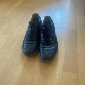 Svarta sneakers från Pantofola d'Oro - Snygga svarta sneakers från Pantofola d'Oro med broderade stjärnor och textdetaljer på sidan och hälen. Skorna har snörning, rund tå och är gjorda i skinn med inslag av mocka. Sulan har en liten detalj i italienska flaggans färger. Perfekta för dig som gillar italiensk stil.