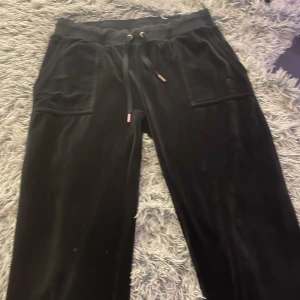 Svarta mjukisbyxor från Juicy Couture - Svarta mjukisbyxor från Juicy Couture i mjuk sammet med bred resår i midjan och snörning. Byxorna har stora fickor fram och bak med knappdetaljer. Perfekta för en chill och trendig look