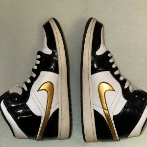 Nike Air Jordans1  i svart, vitt och guld - Snygga Nike Air Jordans i svart, vitt och guld med klassisk snörning. Skorna har en iögonfallande guldfärgad swoosh och detaljer på sidan. Perfekta för den som vill ha en stilren och sportig look.