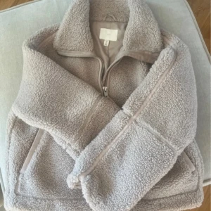 Beige teddyjacka från H&M - Mysig teddyjacka från H&M i beige färg med hög krage och dragkedja framtill. Jackan har två fickor och är tillverkad i fluffigt teddy-material som håller dig varm under kyliga dagar. Perfekt för lager-på-lager och chill höst- och vinterstil.