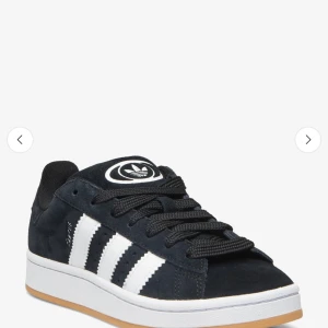 Adidas Campus sneakers svart/vit - Snygga Adidas Campus sneakers i svart mocka med klassiska vita ränder på sidorna och vit sula. Skorna har svart snörning och en gummisula i ljusbrun ton. Perfekta för en streetstyle-look.