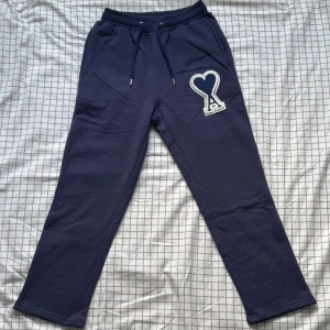 Mörkblå mjukisbyxor från Ami Paris - Mörkblå mjukisbyxor från Ami Paris med snörning i midjan och en stor broderad Ami-hjärtlogga på ena benet. Byxorna är tillverkade i mjuk bomull och har en relaxed passform som är perfekt för chill dagar.