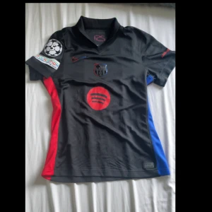 FC Barcelona svart Nike matchtröja - Säljer en svart FC Barcelona matchtröja från Nike med Dri-FIT-material. Tröjan har korta ärmar, klubbmärke i svart med röd och blå kant, röd Spotify-logga på bröstet. Snygga röda och blå detaljer på sidorna.                                                 Passar XS                                                                          Kvitto och Barcelona bag finns, köpt från butiken i Barcelona