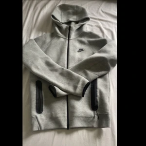 Grå Nike Tech Fleece hoodie med dragkedja - Snygg grå Nike Tech hoodie från Nike med svart logga på bröstet. Tröjan har hel dragkedja, huva och två fickor med svarta detaljer. Perfekt för chill eller träning, och tillverkad i mjuk bomullsblandning för extra komfort.                                      .                                           