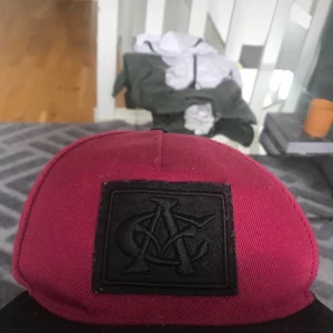 Vinröd keps med svart patch - Snygg vinröd keps med svart patch framtill med broderad logga. Kepsen har klassisk form med svart knapp på toppen och justerbar snapback baktill. Materialet är slitstark bomull och designen är clean och modern.