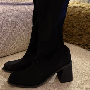 Svarta stretchiga stövletter med klack - Säljer ett par svarta stövletter med hög, bred klack och rund tå. Skorna har en stretchig, strumpliknande överdel som sitter tight runt benet och ger en snygg siluett. Perfekta för dig som gillar minimalistisk och stilren look.