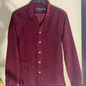 Vinröd manchester-skjorta Ralph Lauren - Snygg vinröd skjorta från Ralph Lauren i mjuk manchester med klassisk krage och slim fit-passform. Långa ärmar, vita knappar framtill och ikonisk blå broderad logga på bröstet. Perfekt för dig som vill ha en stilren och cool look. 