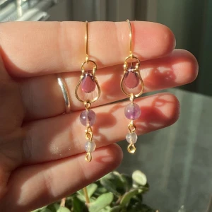 Lila och guldiga örhängen med pärlor - Unika örhängen med droppformad lila pärla i guldinfattning, dekorerade med runda lila och vita pärlor i rad. Hängande design med tunna guldfärgade krokar. Perfekt för dig som gillar färg och detaljer!