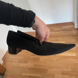 Svarta mocka pumps med spetsig tå - Snygga svarta pumps i mocka med spetsig tå och låg klack. Klassisk och stilren modell med platt sula och rundad öppning. Perfekt för dig som gillar minimalistisk design och vill ha ett par skor som sticker ut med sin form. Storlek 37 från Pancaldi. 