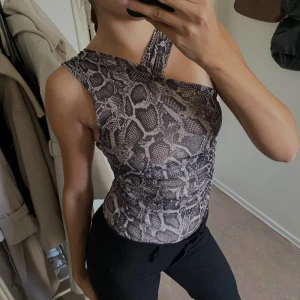 Grå one shoulder topp med ormmönster - Säljer en snygg grå one shoulder topp med ormmönster. Toppen har en asymmetrisk design med breda axelband som korsar framtill och är figurnära. Materialet är stretchigt och mjukt. Helt ny! Aldrig använd. 💗