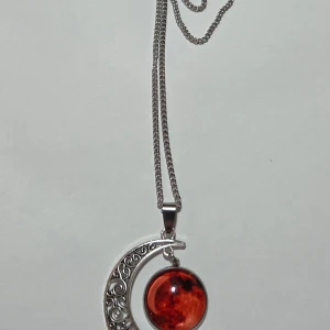 Blood Moon Halvmåne Halsband Självlysande Fullmåne Crescent Glow in Dark 50 cm - Fantastiskt halsband med kosmiskt tema. Hänget är en stor, detaljerad halvmåne i silvertonad metall med filigranmönster, som omfamnar en rund sten.  Den runda stenen har ett mörkt rött/orange Blood Moon-mönster som lyser i mörkret (Glow in the Dark) och avger ett ljust sken efter att ha laddats i ljus.  Detta är det perfekta smycket för den som älskar mystik, astrologi, Wicca eller Gotisk/ Bohemisk stil. Effekt: Självlysande / Glow in the Dark Färg: Silvertonad kedja, Röd/Orange (Blood Moon)