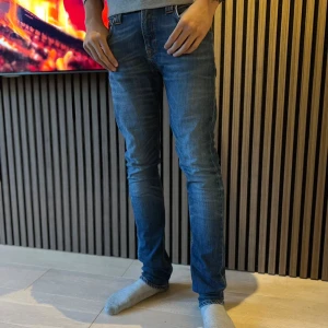 Blå slimfit jeans från Nudie Jeans - Säljer ett par blå slim jeans från Nudie Jeans i storlek W30 L34. Jeansen har klassisk femficksdesign, orangea sömmar och Nudie Jeans signaturbrodyr på bakfickorna. Tillverkade i stretchig denim för en tight passform. Har reparerats i greven och baksidan av baken. Hör av dig vid frågor.
