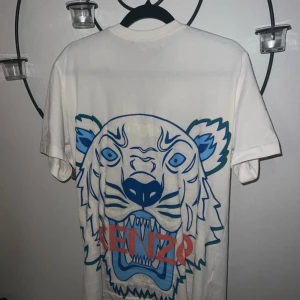 Vit Kenzo t-shirt med tigertryck - Snygg vit t-shirt från Kenzo med stort blått och grönt tigertryck och röd logga på framsidan. Klassisk passform och rund halsringning. Tillverkad i mjuk bomull, perfekt för dig som vill sticka ut med en ikonisk design.