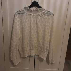 Vit blus med struktur från ONLY - Säljer en vit blus från ONLY med transparent tyg och strukturmönster. Blusen har långärmade ärmar, smock vid hals och nederkant samt volangdetaljer. Materialet är lätt och luftigt, perfekt för dig som gillar romantisk stil.