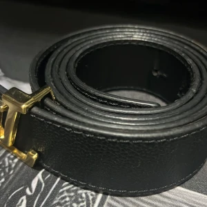 Svart skärp med guldfärgat H-spänne - Snyggt svart bälte med ett stilrent H-format spänne i guldfärg. Bältet är tillverkat i skinn och har en klassisk, slät design som passar till jeans eller kostymbyxor. Perfekt accessoar för att lyfta din outfit med en exklusiv touch.