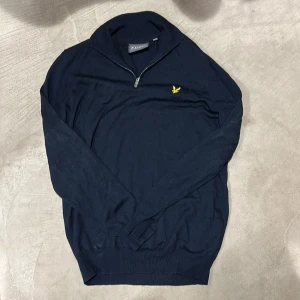 Svart half zip tröja från Lyle & Scott - Snygg svart långärmad tröja från Lyle & Scott med gul broderad logga på bröstet. Tröjan har half zip-dragkedja vid halsen och är stickad i mjukt material. Perfekt för lager-på-lager och stilren look.