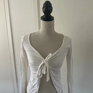 Vit långärmad bolerotopp med knyt - Säljer en vit långärmad bolerotopp med djup v-ringning och knyt framtill. Toppen är croppad och har en smickrande passform som framhäver midjan. Perfekt att styla över ett linne eller klänning för en trendig look.