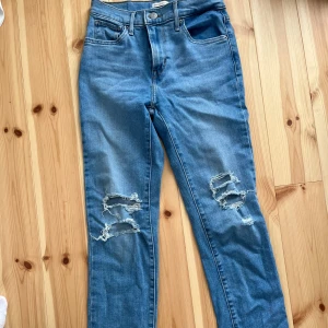 Levi's blå ripped jeans 724 - Snygga blå jeans från Levi's med raka ben. Jeansen har slitna detaljer och hål på båda knäna. Klassisk femficksmodell med Levi's patch bak och röd etikett på fickan.
