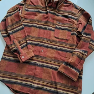 Randig skjorta från Marlboro Classics - Snygg långärmad skjorta från Marlboro Classics i jordnära toner av brun, röd och beige. Skjortan har horisontella ränder, bröstficka med knapp och klassisk krage. Ullmix ger en mjuk och lite rustik känsla. Perfekt för dig som gillar vintagevibbar.