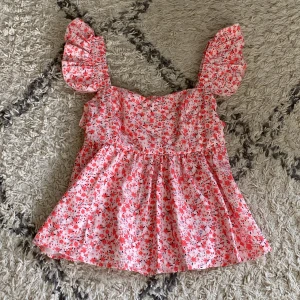 Blommig volangtopp i rosa och rött - Supersöt blommig topp med volangärmar i rosa och rött. Toppen har en luftig passform och ärmlös design med breda axelband och volangdetaljer. Perfekt för varma dagar och ger en romantisk vibe till din outfit.