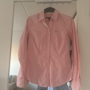 Randig rosa skjorta från GANT - Säljer en klassisk rosa och vit randig skjorta från GANT i storlek 34. Skjortan har lång ärm, knappar framtill och liten broderad logga på bröstet. Tillverkad i 100% bomull för en skön och luftig känsla.