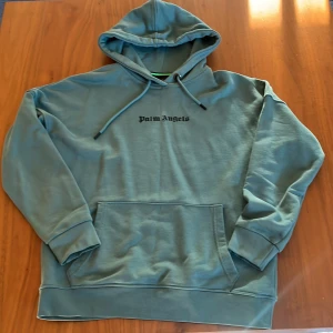 Blå hoodie från Palm Angels - Säljer en blå hoodie från Palm Angels med stor svart logga på ryggen och mindre logga framtill. Tröjan har huva med snörning och en stor magficka. Skön och avslappnad passform, perfekt för chill dagar.