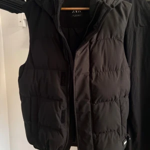 Svart dunväst med huva från Zara - Svart dunväst från Zara med quiltad design och praktisk huva. Västen har dragkedja framtill och två fickor på sidorna. Perfekt för lager-på-lager och en clean look. Passar dig som gillar stilren och funktionell streetwear.