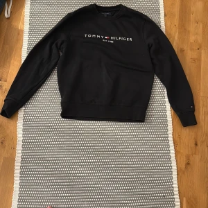 Svart sweatshirt från Tommy Hilfiger - Svart sweatshirt från Tommy Hilfiger med broderad logga och text på bröstet. Klassisk rund halsringning, ribbade muddar och mjuk insida. Perfekt för en chill och stilren look. Passar dig som gillar enkel och tidlös design.