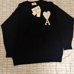 Svart stickad tröja från Ami Paris - Svart stickad tröja från Ami Paris med vit hjärtlogga och bokstaven A på bröstet. Klassisk rund halsringning och långa ärmar med ribbade muddar. Perfekt för dig som gillar stilrena och trendiga plagg.