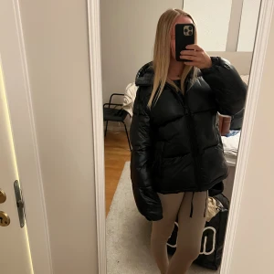 Svart pufferjacka med huva - Säljer en svart pufferjacka med huva och dragkedja framtill. Jackan har en oversized passform och är perfekt för kalla dagar. Den är tillverkad i ett glansigt syntetmaterial och har långa ärmar. Snygg och trendig stil som passar till många outfits.