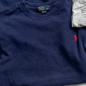 Marinblå t-shirt från Polo Ralph Lauren - Snygg marinblå t-shirt från Polo Ralph Lauren med klassisk röd logga broderad på bröstet. T-shirten har rund halsringning och är kortärmad, tillverkad i mjuk bomull för en skön känsla. Barnstorlek passar S-xs 