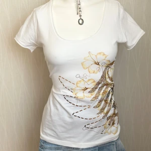 Vit t-shirt med blommor och paljetter - Vit t-shirt från Jolokia med korta ärmar och rund halsringning. Framsidan har ett stort blommönster i beige och brunt, dekorerat med glittrande paljetter. T-shirten är i mjuk bomull och har en feminin, figurnära passform.