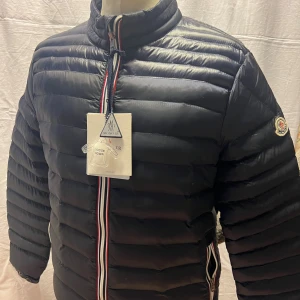 Svart Moncler dunjacka herr - Svart dunjacka från Moncler med klassisk quiltad design och Moncler-logga på ärmen. Jackan har dragkedja framtill med röd, vit och blå detalj samt hög krage. Fylld med gåsdun för extra värme och stilren look. Perfekt för kalla dagar.