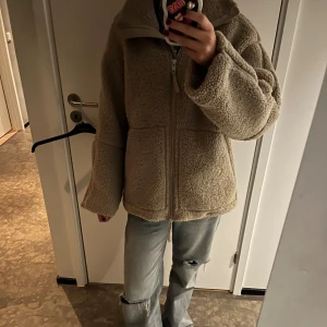 Beige teddyjacka med dragkedja - Mysig teddyjacka i beige med hög krage och hel dragkedja framtill. Jackan har två stora fickor och en loose passform som ger en chill vibe. Perfekt för kalla dagar.