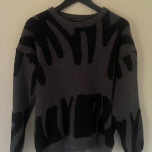 Grå och svart stickad tröja med mönster - Säljer en grå stickad tröja med svart abstrakt mönster. Tröjan har rund halsringning och långa ärmar. Den är oversized och har en cool, konstnärlig vibe som sticker ut. Perfekt för dig som gillar unika plagg och vill ha något extra i garderoben.