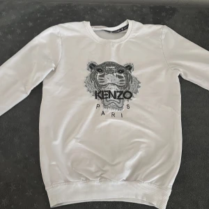 Vit Kenzo sweatshirt - Säljer en vit sweatshirt från Kenzo med ikonisk tigerbrodyr och texten 'Kenzo Paris' framtill. Tröjan har rund hals, långa ärmar och ribbade muddar. Perfekt statement-plagg för dig som gillar streetstyle och vill sticka ut.