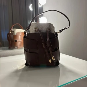 Brun bucket handväska i skinn och canvas - Snygg bucket handväska i mörkbrunt skinn med detaljer i beige canvas. Väskan har ett coolt flätat mönster och stängs med snörning upptill. Guldiga metalldetaljer och kort handtag gör den både trendig och praktisk. Perfekt för dig som gillar stilrena accessoarer.