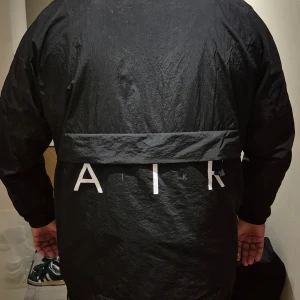 Svart Nike Air vindjacka - Säljer en svart Nike Air vindjacka med hög krage och hel dragkedja framtill. Jackan har elastiska muddar vid ärmslut och stor AIR-logga på ryggen. Materialet är lätt och syntetiskt, perfekt för att hålla vinden ute. Snygg och sportig stil med diskret Nike Air-märke på bröstet. Gör att göra till en tygkasse. 