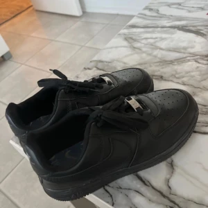 Nike Air Force 1 helsvarta sneakers - Säljer ett par klassiska Nike Air Force 1 i helsvart skinn. Skorna har perforerad tå, svart sula och svarta skosnören med silverfärgade dubraes. Perfekta för dig som vill ha en clean och tidlös look. OBS INTE ÄKTA
