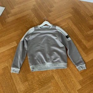 Grå sweatshirt från Stone Island - Säljer en stilren grå sweatshirt från Stone Island med klassisk logga på ärmen. Tröjan har rund halsringning, ribbade muddar och kontrasterande paneler på axlar och ärmar. Perfekt för dig som gillar streetwear och snygga detaljer.