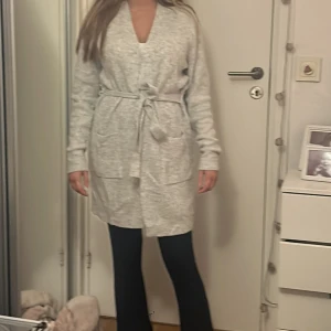 Ljusgrå lång kofta med knytband - Mysig ljusgrå lång kofta med knytband i midjan och stora fickor framtill. Koftan har v-ringning och långa ärmar, perfekt att slänga på sig över en t-shirt eller topp. Enkel och stilren design som funkar året runt.