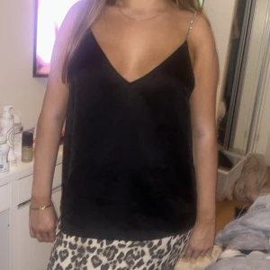 Svart linnetopp med glittriga band - Säljer en svart linnetopp med glansigt material och smala glittriga axelband. Toppen har v-ringning både fram och bak och är ärmlös. Perfekt för dig som vill ha en enkel men snygg topp med lite extra bling.