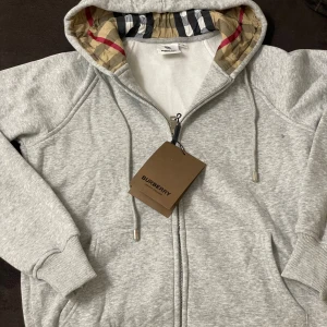 Grå hoodie från Burberry zipper - Snygg grå hoodie från Burberry med klassiskt rutigt foder i huvan och dragkedja framtill. Tröjan har ribbade muddar, fickor och snörning i huvan. Perfekt för dig som vill ha en stilren och exklusiv look.