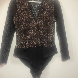 Svart spetsbody med blommönster H&M - Svart body från H&M i storlek 38, med långärmade spetsärmar och blommigt spetsmönster över hela toppen. V-ringad framtill och med knäppning i grenen. Materialet är mjuk spets och syntet, perfekt för att ge en cool och elegant vibe.