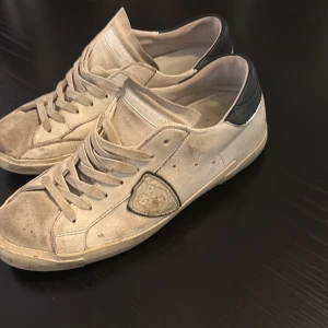 Philippe Model vita sneakers skinn - Säljer ett par vita Philippe Model sneakers i skinn med svart häl och klassisk logotyp på sidan. Skorna har rund tå, platt sula och snörning framtill. Stilren design med diskreta sömmar och detaljer, perfekt för dig som gillar exklusiva sneakers. Kan gå ner pris vid snabb affär