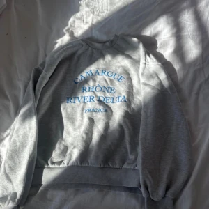 Grå sweatshirt från Gina Tricot - Grå sweatshirt från Gina Tricot med blå texttryck 'Camargue Rhône River Delta France' på bröstet. Tröjan har rund hals, långa ärmar och ribbade muddar vid ärmslut och nederkant. Perfekt för en chill och avslappnad stil. I storlek S