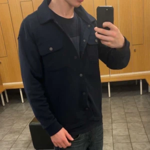 Mörkblå overshirt med tryckknappar - Säljer en stilren mörkblå overshirt. Materialet är tunnare, mjukt och är troligtvis bomull eller polyester. Knappt använd så nytt skick! Inga defekter alls. 