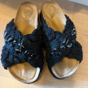 Tofflor - Snygga svarta sandaler från skopunktrn med breda korslagda band i stickad textil och dekorerade med svarta pärlor och stenar. Sandalerna har öppen tå och en mjuk, beige innersula som ger skön komfort. Perfekt för dig som gillar detaljer och vill sticka ut. Nypris 449kr