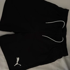 Svarta shorts från Puma - Svarta sportiga shorts från Puma med vit logga på benet och vit snörning i midjan. Perfekta för träning eller chill, tillverkade i ett lätt och bekvämt syntetmaterial. Klassisk loose fit och elastisk midja för extra komfort.